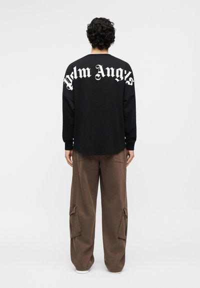 Persona con capelli ricci scuri che indossa una maglia nera a maniche lunghe con la scritta "Palm Angels" sul retro e pantaloni cargo marroni, vista di spalle.