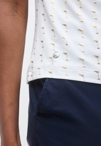 Λευκό πουκάμισο με μικρά χρυσά κεντημένα σχέδια και ασημένιο στρογγυλό λογότυπο κοντά στο στρίφωμα, σε συνδυασμό με σκούρο μπλε παντελόνι.