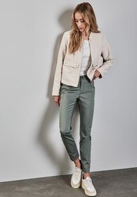 Giacca cropped color beige chiaro con due tasche frontali, abbinata a pantaloni aderenti verdi oliva. Una camicia bianca e sneakers chiare completano l'outfit.