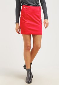 Mini-jupe rouge en tissu lisse, dotée de poches latérales et d'accents de boutons. Associée à un haut gris à manches longues et des bottines noires.