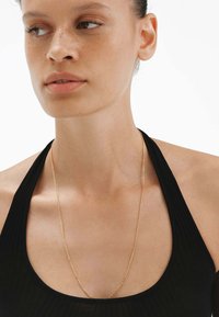 Collar de cadena de metal dorado que se despliega alrededor del cuello, con un diseño fino y delicado. La modelo lleva una camiseta de tirantes negra con canalé.