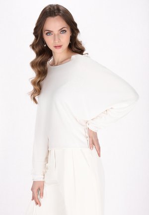 Haut blanc à manches longues avec une coupe ample, présentant un tissu texturé et des accents à cordon aux poignets. Associé à un pantalon blanc plissé.