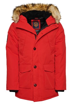 Parka rossa isolata con cappuccio foderato in pelliccia, due tasche anteriori, bottoni a pressione e un patch con logo sulla manica. Tessuto testurizzato.
