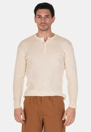 Uomo che indossa un maglione henley beige a maniche lunghe e pantaloni marroni, in piedi dritto davanti a uno sfondo bianco uniforme.