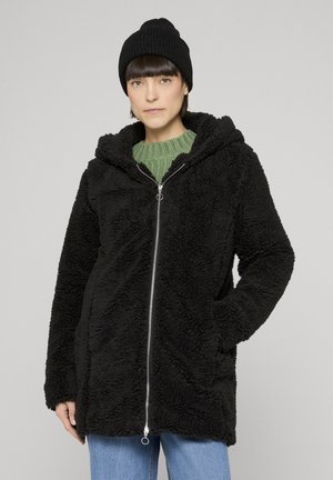 Frau in einer schwarzen, flauschigen Jacke mit Reißverschluss, grünem Strickpullover, schwarzer Mütze und blauen Jeans, die mit einer Hand in der Tasche steht.