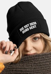 Black knit beanie with ribbed texture and a rectangular patch featuring white text “DIE MIT DEM HUND GEHT.” Warm, casual winter accessory.