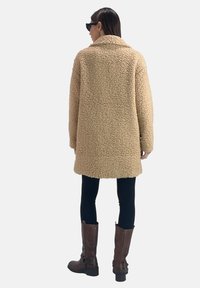 Cappotto beige morbido con colletto, realizzato in tessuto strutturato, con maniche lunghe e che arriva a metà coscia, abbinato a leggings neri e stivali marroni.