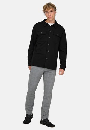 Giovane uomo che indossa una camicia nera con bottoni e tasche con patta, pantaloni grigi a quadri e sneakers nere, in piedi davanti a uno sfondo bianco.