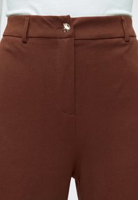 Pantalon marron taille haute avec passants pour ceinture et une seule fermeture par bouton doré brillant à la taille, porté avec un top blanc.