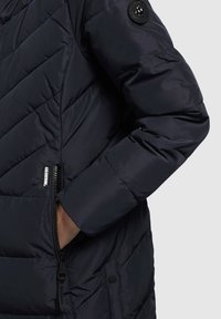 Námornícky pufový kabát s chevronovým prešívaním, bočnou vreckom so zipsom a okrúhlym čiernym logom na rukáve. Elegantný, zateplený dizajn.