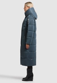 Toppatakki, jossa on korkea kaulus ja huppu, vetoketjulliset taskut sekä kolmionmuotoinen merkkilogo helman lähellä. Yhdistetty mustien kiilakorkokenkien kanssa.