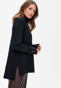 Kobieta z luźnymi falującymi włosami, ubrana w czarny sweter z golfem i brązowe skórzane spodnie, stojąca w profilu na białym tle.