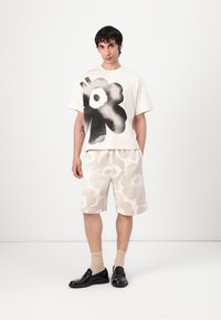 Vit bomullströja med ett stort svart blommönster, matchad med beige shorts som har ett ljusare blommönster. Svarta loafers och strumpor fulländar looken.