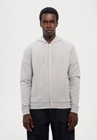 Grå zip-up hettegenser med snorhette, frontlommer og ribbede mansjetter. Myk stoff med subtil tekstur, paret med mørke jeans.