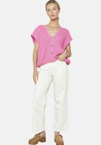 Pītā rozā īsām piedurknēm blūze ar V formas kakla izgriezumu un zelta pogām, apvienota ar baltām platām biksēm un brūnām koka kurpēm. Gluda tekstūra, ikdienišķs dizains.