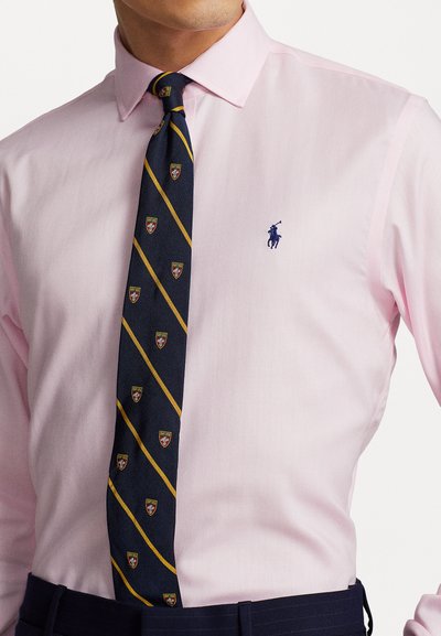 Svijetlo ružičasta košulja s dugim rukavima i herringbone teksturom, s plavim polo logotipom. U paru s tamnoplavim kravatom, koja prikazuje zlatne pruge i embleme.