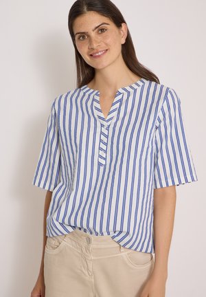 Jeune femme aux longs cheveux foncés, portant une chemise blanche à rayures verticales bleues et un pantalon beige clair, souriant sur un fond neutre.