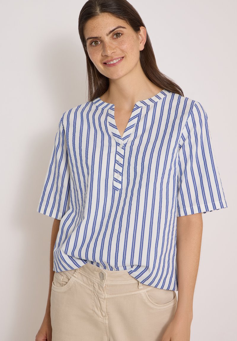 Jeune femme aux longs cheveux foncés, portant une chemise blanche à rayures verticales bleues et un pantalon beige clair, souriant sur un fond neutre.