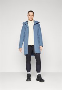 Blå, vanntett jakke med hette, glidelås foran og sidelommer. Kombinert med en kremfarget fleece og svarte tights og fottøy.