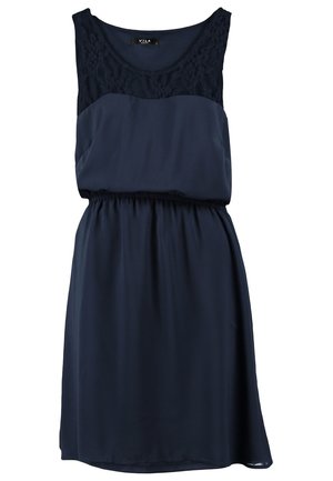 Femme portant une robe sans manches bleu marine avec un col en dentelle, tenant une pochette beige et portant des sandales à talons hauts beiges.