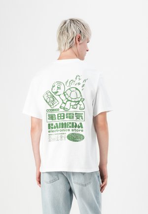 Person with short blonde hair wearing white T-shirt with green turtle holding a cracked phone graphic and text "KAMEDA electronics store" on back.