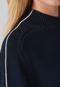 Pull en tricot navy avec un col haut à côtes, présentant des rayures latérales blanches et un tissu texturé. Vue en gros plan mettant en valeur les détails de couture.