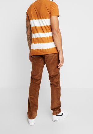 Mand iført orange og hvidstribet T-shirt, brune kordfløjlsbukser, hvide sneakers, stående med ryggen til med synlig tatoveret højre arm.