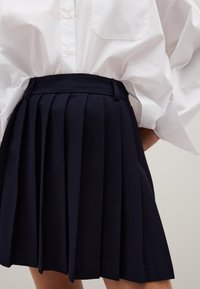 Falda plisada azul marino hecha de tejido entrelazado. Presenta una cinturilla alta y una textura suave. Combinada con una camisa blanca de botones extragrande.