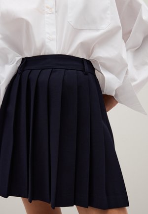 Falda plisada azul marino hecha de tejido entrelazado. Presenta una cinturilla alta y una textura suave. Combinada con una camisa blanca de botones extragrande.