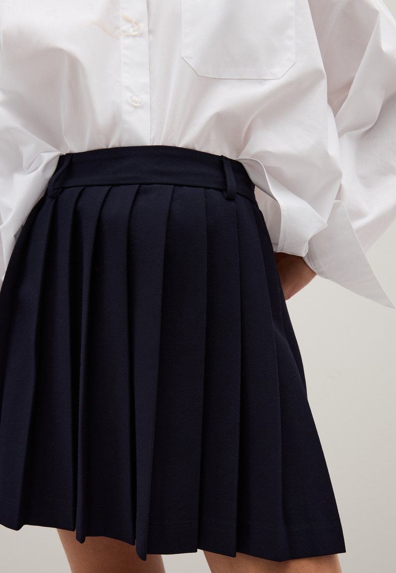 Falda plisada azul marino hecha de tejido entrelazado. Presenta una cinturilla alta y una textura suave. Combinada con una camisa blanca de botones extragrande.