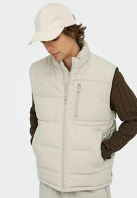 Beige puffer vest med høj krave, lynlåsåbning foran og sidelommer. Bæret over en brun kabelstriktrøje med en glat struktur.