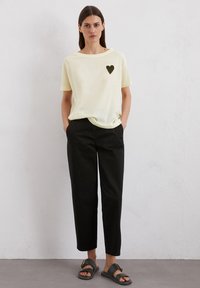 Lichtgele katoenen T-shirt met een zwarte hartafbeelding op de linkerborst, gecombineerd met zwarte loszittende broek en zwarte sandalen.