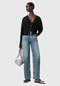 Zwarte cardigan met knoopaccenten, gecombineerd met lichtblauwe brede jeans. Model houdt een zilveren handtas vast en draagt metallic sandalen.