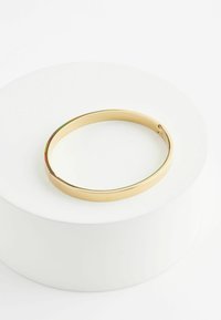 Bracelet en métal doré avec une surface lisse et réfléchissante. Présente un design minimaliste et un fermoir sécurisé. Présenté sur un piédestal circulaire blanc.