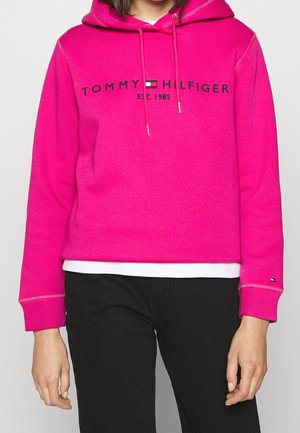 Sweat à capuche Tommy Hilfiger rose vif avec logo et texte "Est. 1985", porté avec un pantalon noir et une chemise blanche qui dépasse en dessous.