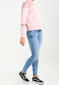 Ljusrosa sweatshirt med spetsdetaljer vid axlarna, ihopparat med tajta ljusblå jeans och svarta sneakers med vita sulor.