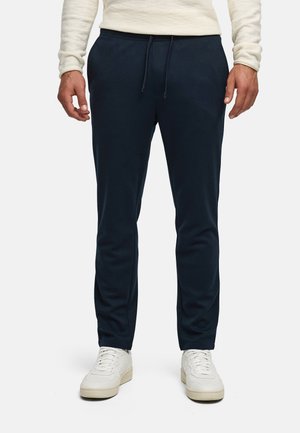 Mężczyzna w granatowych spodniach z troczkami i białych trampkach, stojący na jasnym, jednolitym tle.