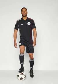 Črn komplet polo majice in kratkih hlač s rdečimi poudarki, z logotipom Adidas in okroglim logotipom. Model stoji na nogometni žogi z črnimi kopačkami.