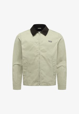 Veste beige à fermeture éclair avec col noir, deux poches avant et une petite étiquette de marque rectangulaire sur le côté gauche de la poitrine.