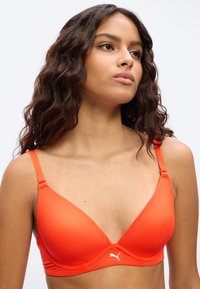 Kvinne med langt krøllete hår som bærer en lys oransje Puma sportstopp, ser lett oppover og til siden mot en lys bakgrunn.