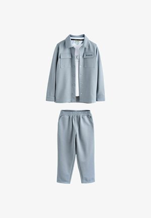 Svetlomodra bunda na gumbe z dvema žepoma na prsih in ujemajoče se elastične hlače z visokim pasom, kombinirana z belo tiskano majico spodaj.