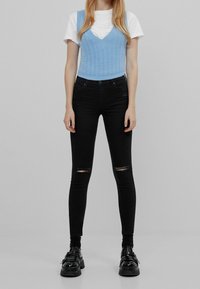 Top corto de punto azul claro sobre una camiseta blanca, combinado con jeans skinny negros desgastados y zapatos negros robustos con hebillas.