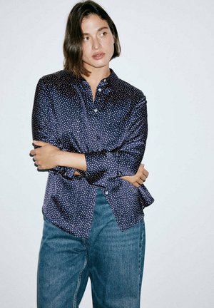 Femme aux cheveux courts bruns portant une chemise bleu marine à pois et un jean bleu, debout, les bras croisés devant un fond blanc.