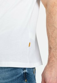 Hvid bomulds t-shirt med korte ærmer, en lige kant og et lille gult mærke på venstre side. Båret med blå denim jeans.