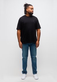 Άνδρας με dreadlocks που φοράει μαύρο μπλουζάκι, μπλε τζιν και λευκά αθλητικά παπούτσια, στέκεται και κοιτάζει προς τα δεξιά πάνω σε ένα απλό φόντο.