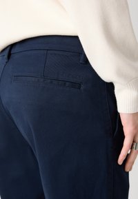 Navy blue pants made from a smooth fabric, featuring a back pocket, subtle branding, and a tailored fit. Textured sweater visible above.