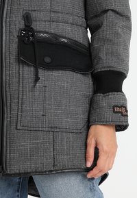 Grå vadderad jacka med svarta detaljer, som har en dragkedjeficka, elastiska ärmmuddar och en logotypapplikation. Strukturerad tyg med rutigt mönster.