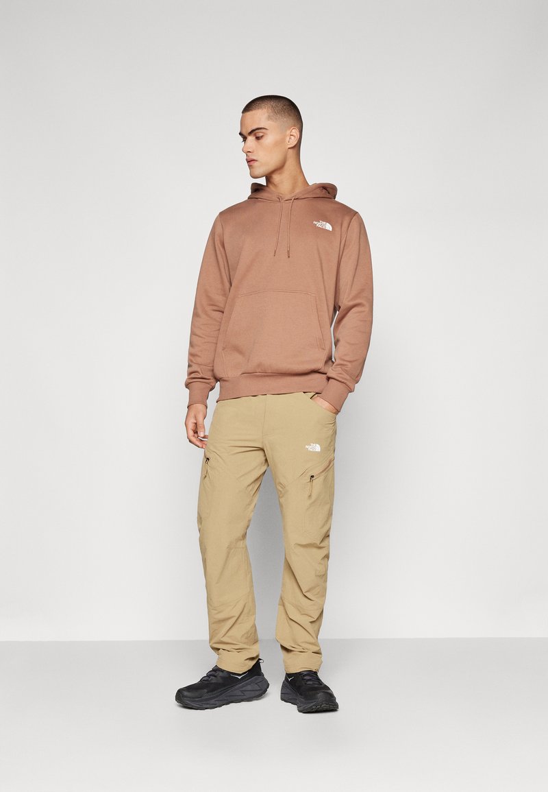 Brauner Kapuzenpullover mit einer Vordertasche; beige Cargo-Hosen mit seitlichen Reißverschlüssen; schwarze Schuhe. Minimalistische Logoanpassungen auf beiden.
