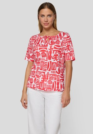 Frau trägt ein weißes Kurzarmshirt mit roten abstrakten Formen und weiße Hose, steht vor einem einfarbigen hellgrauen Hintergrund.