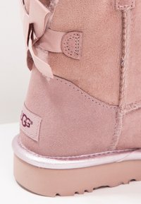 Růžová kotníková bota z semiše s detailem saténové mašle, plyšovou podšívkou a metalickou růžovou podrážkou. Na boku je logo UGG.